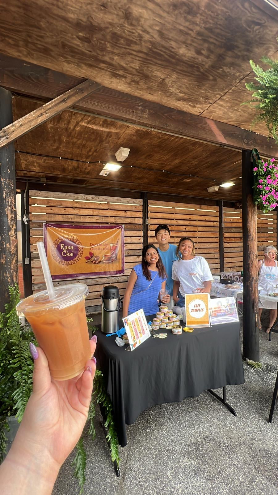 A group of 3 people at the University City Farmers Market stand behind a table, which has a pile of masala chai tins and a sign that says "free samples". Behind them is a bright yellow and maroon banner that says "Rasa Chai." In the front of the photo, a hand with manicured purple nails holds a transparent cup of iced masala chai.