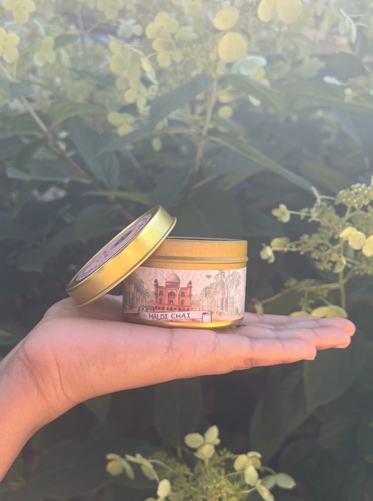 A golden tin of chai with a label depicting a traditional Indian style building and a white scroll labeled Haldi Chai. In the background is a bed of colorful yellow flowers.