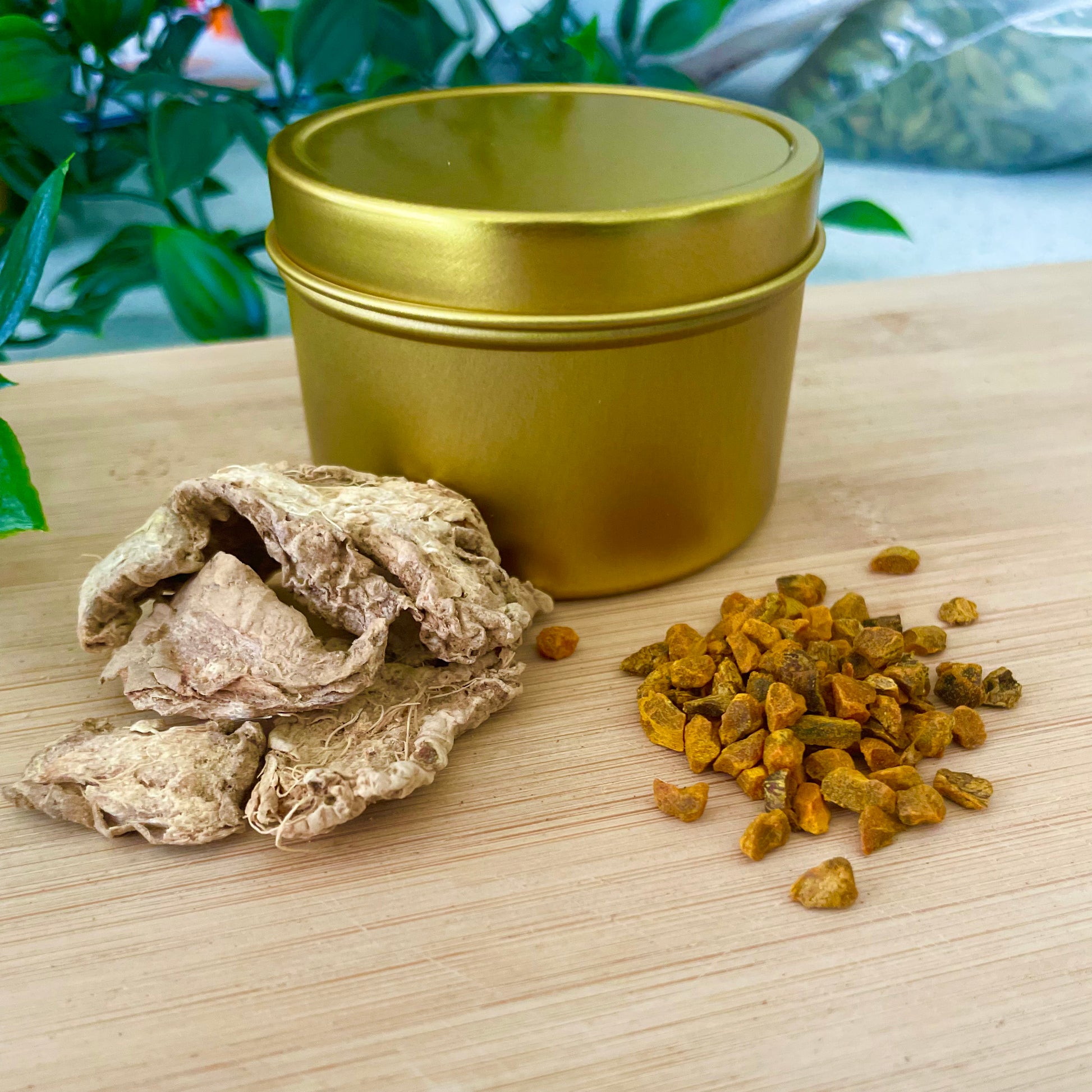 A blank golden tin sitting on a wooden counter. A pinch of dried ginger and bright orange dried turmeric sit in front of the tin.