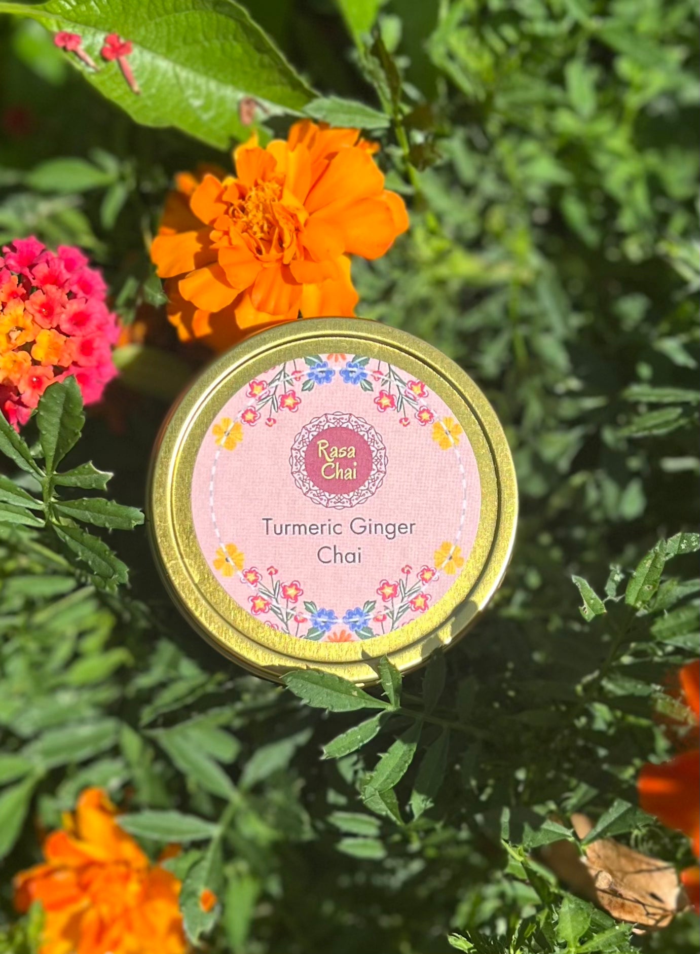 A golden tin of chi sitting in a bed of vibrant orange, pink, and yellow flowers. The tin is labeled "Rasa Chai Turmeric Ginger Chai"