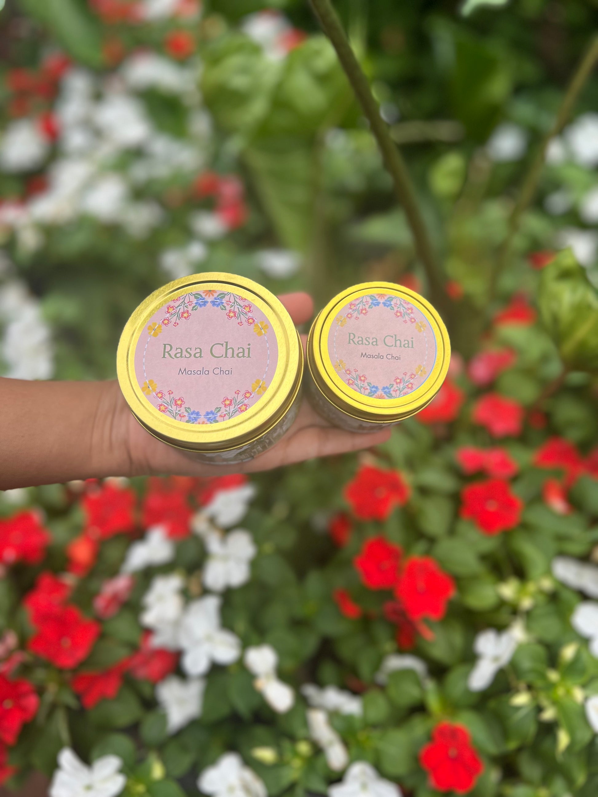 Hand extended with a small and large tin of Rasa Chai masala chai with a bush of colorful flowers in the background