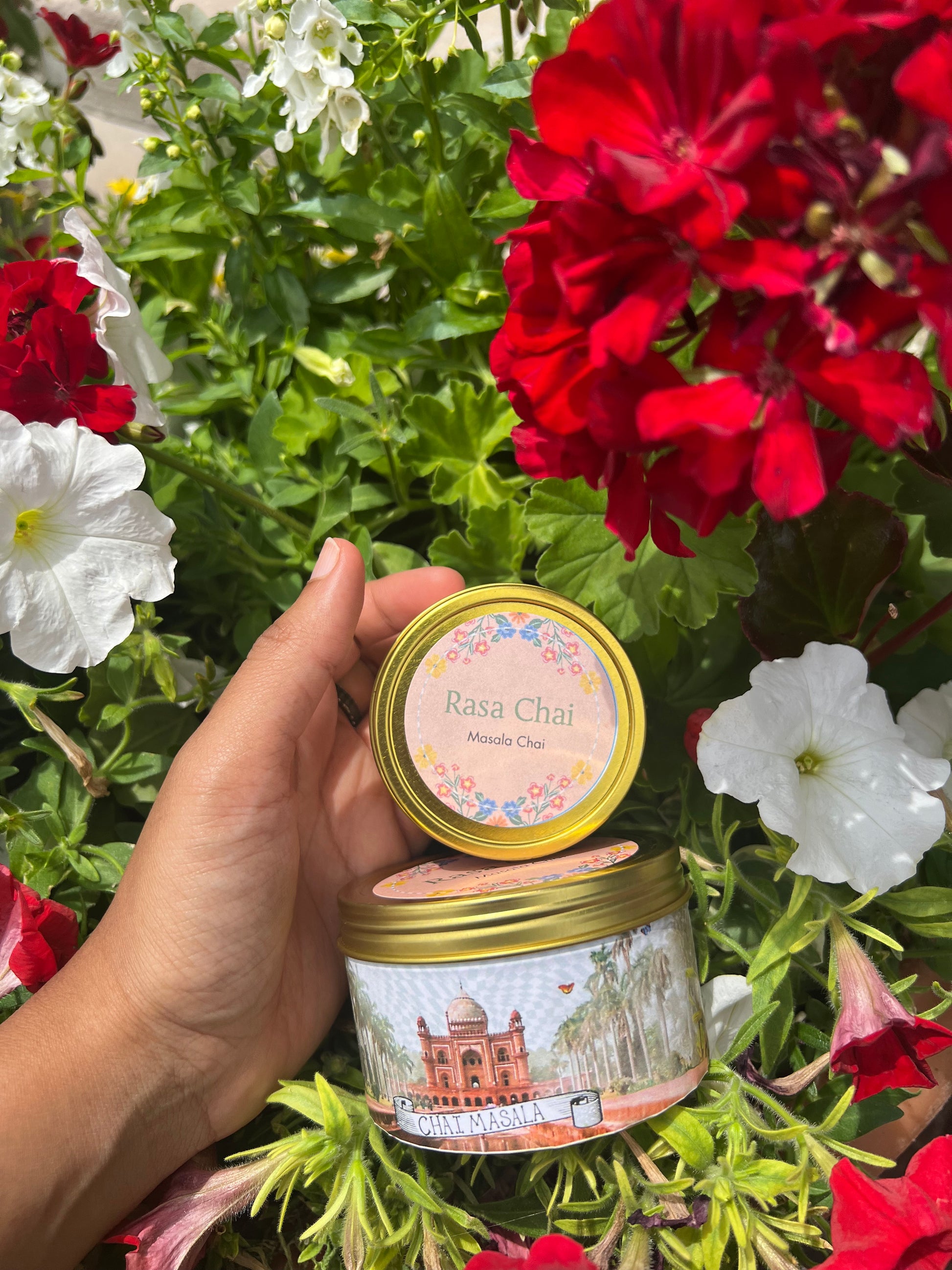 Hand next to a small and large tin of Rasa Chai masala chai with a bush of colorful flowers in the background