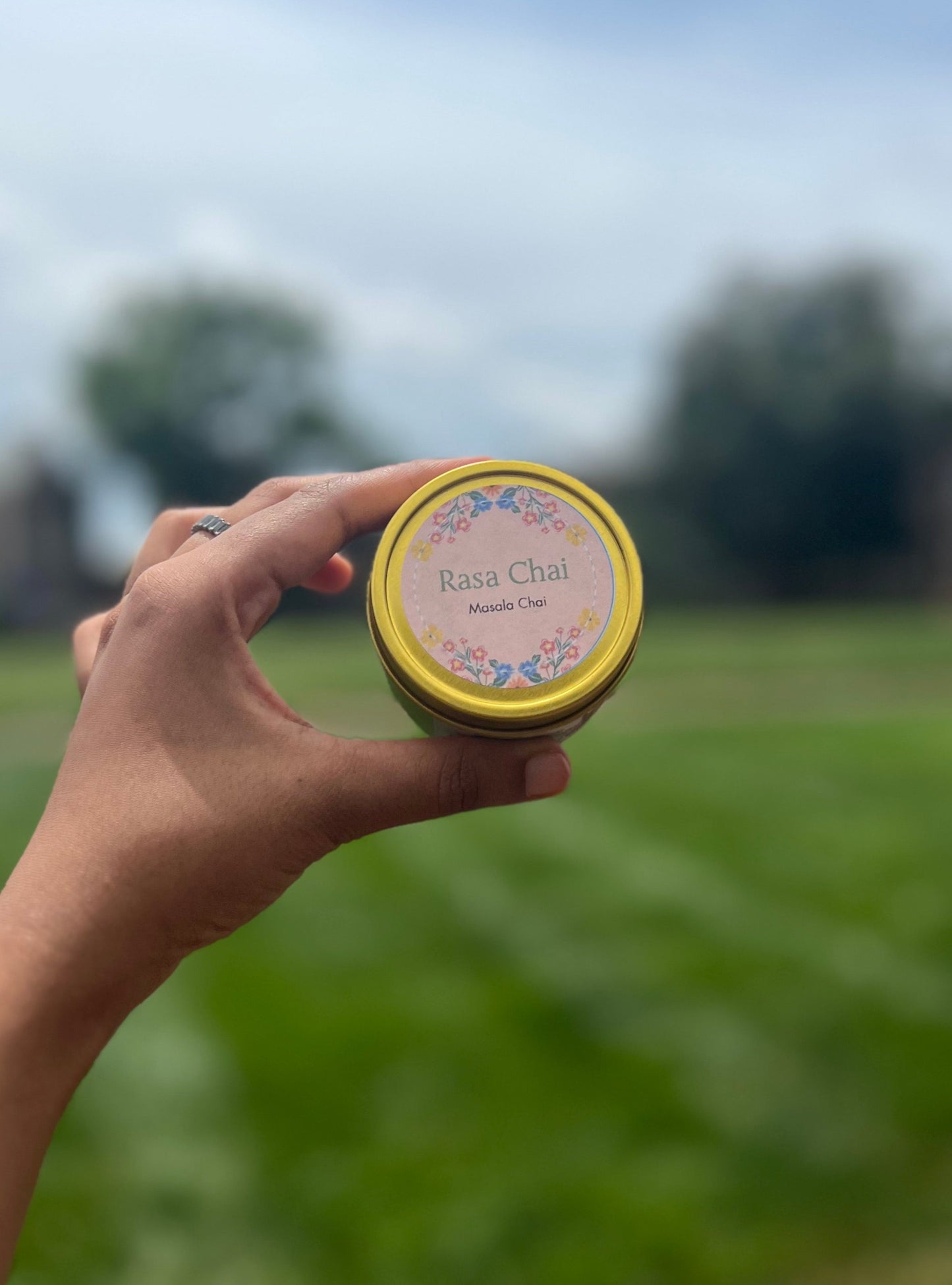 Hand holding a tin of "Rasa Chai" masala chai in front of an open field