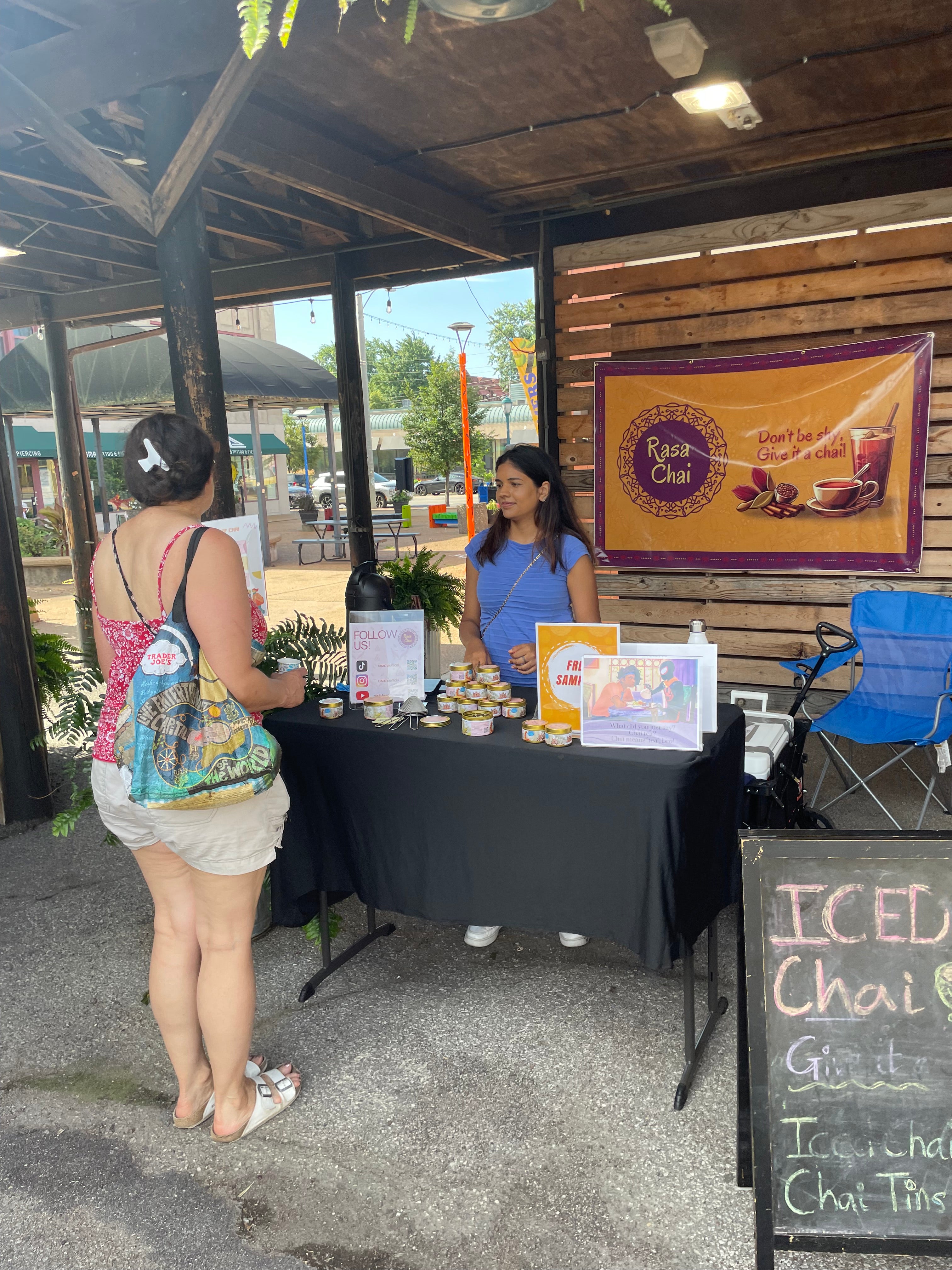 A woman in a pink tanktop and shorts stands before a desk of masala chai tins as another woman in a blue shirt speaks to her. Behind the woman in the blue shirt is a clorful yellow and maroon banner that reads, "Rasa Chai". On the side of the table is a chalkboard that says "iced chai"