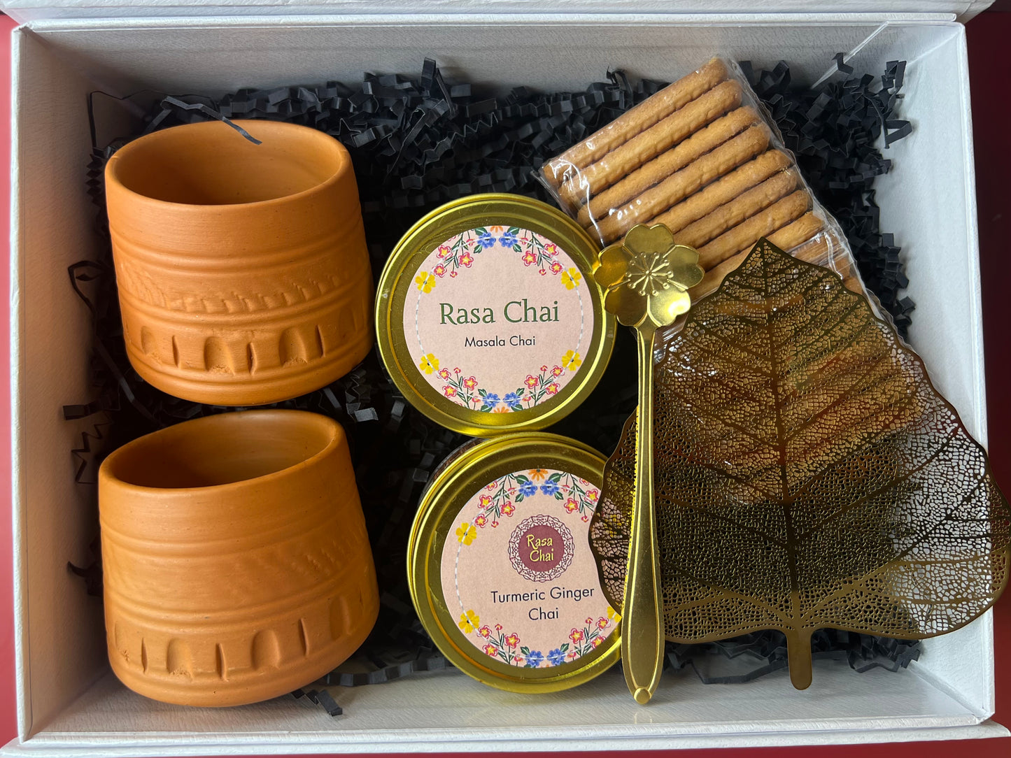 A white box containing two clay kulhads, 21 golden tin labeled "Rasa Chai Masala Chai", another tin labeled "Rasa Chai Turmeric Ginger Chai", A golden spoon in the shape of a flower, a sleeve of Parle G biscuits, and a golden leaf-shaped strainer.