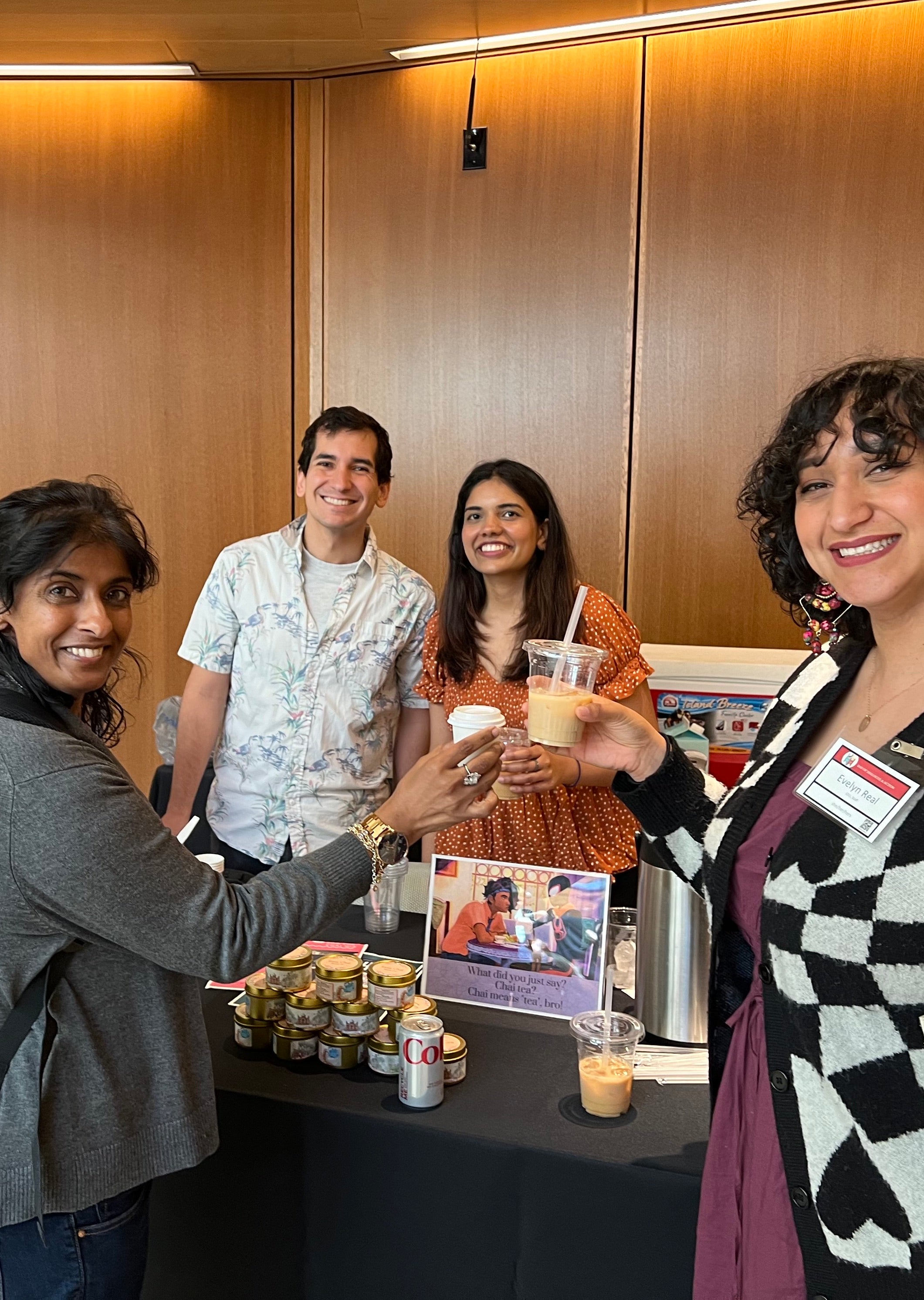 A group of four people gathered around a table, smiling as they hold cups of iced and hot masala chai. On the table is a sign featuring the "chai tea" scene from Spider Man: Into the Spiderverse, a pile of masala chai tins, and a silver can of soda.